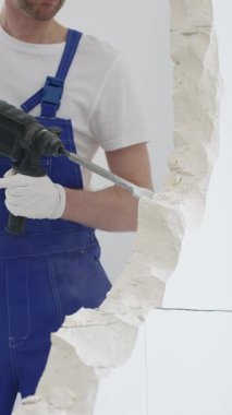 Mavi tulumlu ve koruyucu beyaz eldivenli tanımlanamayan erkek inşaat işçisi, beyaz duvarı döner çekiç matkapıyla yıkıyor, toz üretiyor ve yatay görüşü kapatıyor. Yenileme