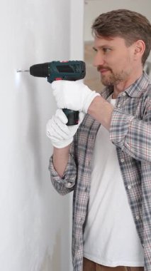 Gri gömlekli ve beyaz eldivenli orta yaşlı işçi, ev yenileme çalışmaları sırasında kablosuz matkapla duvarda delik açıyor. Dikey görünüm.