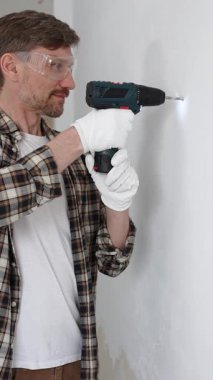 İşaretli gömlek ve beyaz eldiven giyen orta yaşlı bir adam, ev yenileme çalışmaları sırasında kablosuz matkapla duvarda delik açıyor. Dikey görünüm.
