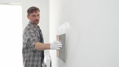 Koruyucu eldiven ve ekose gömlek giyen erkek inşaat işçisi, tadilat projesi sırasında profesyonel alçı levha bantlama bıçağı kullanarak duvarın yüzeyine sorunsuzca alçı yayıyor. Dikey görünüm.