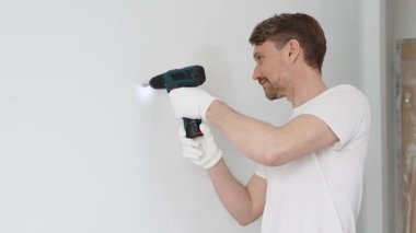 Beyaz tişört ve koruyucu eldiven giyen orta yaşlı erkek işçi, ev yenileme çalışmaları sırasında kablosuz matkapla duvarda delik açıyor. Dikey görünüm.