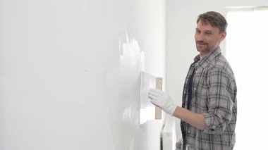 Koruyucu eldiven ve ekose gömlek giyen erkek inşaat işçisi, tadilat projesi sırasında profesyonel alçı levha bantlama bıçağı kullanarak duvarın yüzeyine sorunsuzca alçı yayıyor. Dikey görünüm.