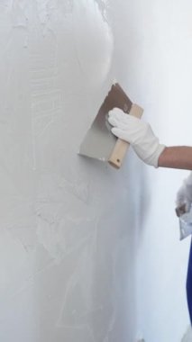 Koruyucu eldivenler ve mavi inşaat tulumları giyen erkek inşaat işçisi, tadilat projesi sırasında profesyonel alçı levha bantlı bıçak kullanarak duvarlara sorunsuzca alçı yayıyor. Portre