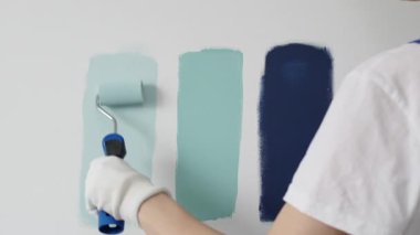 Eldiven giyen ressam beyaz bir duvara boya silindiri uyguluyor, mavi rengin çeşitli tonlarını test ederek iç tasarım için güzel bir eğim etkisi yaratıyor.