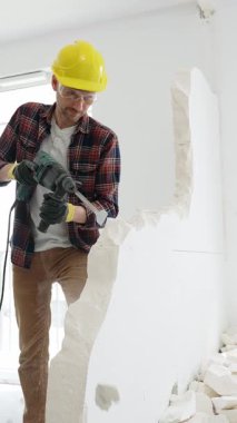 Bej kareli gömlek, sarı başlık ve koruyucu eldiven giyen inşaat işçisi, dönen çekiç matkabıyla beyaz duvarı yıkıyor ve orta uzunlukta toz üretiyor. Yenileme konsepti