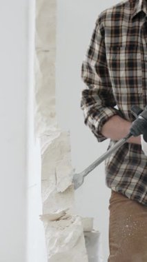 Tanınmayan bej kareli gömlek ve koruyucu eldivenler giyen erkek inşaat işçisi, döner çekiç matkabıyla beyaz duvarı yıkıyor, toz üretiyor, yakın görüş sağlıyor. Yenileme konsepti.