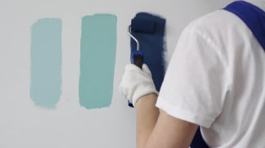 Eldiven giyen ressam beyaz bir duvara boya silindiri uyguluyor, mavi rengin çeşitli tonlarını test ederek iç tasarım için güzel bir eğim etkisi yaratıyor.