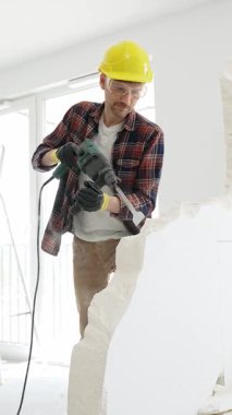 Bej kareli gömlek, sarı başlık ve koruyucu eldiven giyen inşaat işçisi, dönen çekiç matkabıyla beyaz duvarı yıkıyor ve orta uzunlukta toz üretiyor. Yenileme konsepti