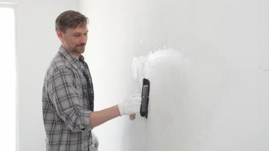 Eldiven ve ekose gömlek giyen orta yaşlı inşaat işçisi, yenileme projesi sırasında pürüzsüz bir yüzey oluşturmak için beyaz duvar alçılamak için son malayı kullanıyor..