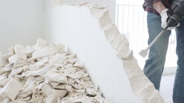 Kırmızı kareli kotlar ve koruyucu eldivenler giyen kimliği belirsiz erkek inşaat işçisi, döner çekiç matkabıyla beyaz duvarı yıkıyor, yerde enkaz oluşturuyor, manzarayı kapatıyor. Yenileme.