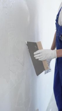 Koruyucu eldivenler ve mavi inşaat tulumları giyen erkek inşaat işçisi, tadilat projesi sırasında profesyonel alçı levha bantlı bıçak kullanarak duvarlara sorunsuzca alçı yayıyor. Portre