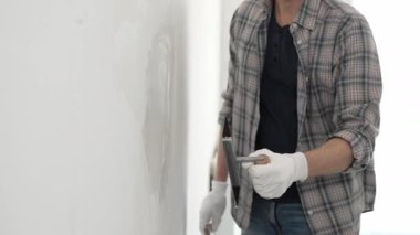 Eldiven ve ekose gömlek giyen kimliği belirsiz erkek inşaat işçisi, yenileme projesi sırasında pürüzsüz bir yüzey oluşturmak için beyaz bir duvar alçılamak için son malayı kullanıyor..