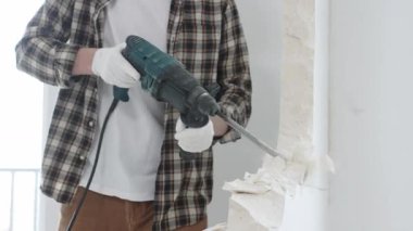 Tanınmayan bej kareli gömlek ve koruyucu eldivenler giyen erkek inşaat işçisi, döner çekiç matkabıyla beyaz duvarı yıkıyor, toz üretiyor, yakın görüş sağlıyor. Yenileme konsepti.