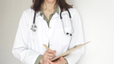 Yeşil bir bluzun üzerine steteskop ve sağlık önlüğü giyen kimliği belirsiz kadın doktor, beyaz bir panoya tıbbi uzmanlığı ve hasta bakımını temsil eden bir yazı yazıyor. Tıp konsepti.