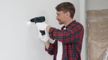 Kırmızı tişört ve beyaz eldiven giyen orta yaşlı erkek işçi, ev yenileme çalışmaları sırasında kablosuz matkapla duvarda delik açıyor. Dikey görünüm.