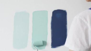 Eldiven giyen ressam beyaz bir duvara boya silindiri uyguluyor, mavi rengin çeşitli tonlarını test ederek iç tasarım için güzel bir eğim etkisi yaratıyor.