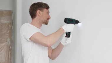 Beyaz tişört ve koruyucu eldiven giyen orta yaşlı erkek işçi, ev yenileme çalışmaları sırasında kablosuz matkapla duvarda delik açıyor. Dikey görünüm.