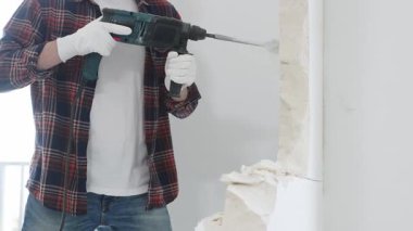 Kırmızı kareli kot pantolon ve koruyucu eldiven giyen kimliği belirsiz erkek inşaat işçisi, döner çekiç matkabıyla beyaz duvarı yıkıyor, toz üretiyor, yakın görüş sağlıyor. Yenileme konsepti.