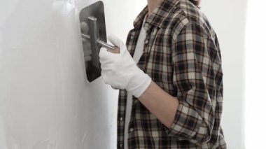 Koruyucu eldiven ve kareli tişört giyen kimliği belirsiz kadın inşaat işçisi, beyaz duvarda ıslak alçıyı düzleştiriyor ve iç dekorasyon sırasında profesyonel malayla malzeme yayıyor.