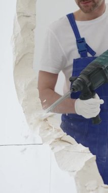 Mavi tulumlu ve koruyucu beyaz eldivenli tanımlanamayan erkek inşaat işçisi, beyaz duvarı döner çekiç matkapıyla yıkıyor, toz üretiyor ve yatay görüşü kapatıyor. Yenileme
