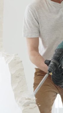 Beyaz tişörtlü ve koruyucu eldivenli, kimliği belirsiz bir inşaat işçisi, beyaz duvarı döner çekiç matkapıyla yıkıyor, yerde enkaz oluşturuyor, dikey yakın görüş açısında. Yenileme.