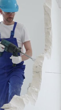 Mavi tulumlu ve koruyucu beyaz eldivenli bir inşaat işçisi, beyaz duvarı döner çekiç matkabıyla yıkıyor, toz üretiyor, yatay görüşü kapatıyor. Yenileme konsepti.