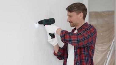 Kırmızı tişört ve beyaz eldiven giyen orta yaşlı erkek işçi, ev yenileme çalışmaları sırasında kablosuz matkapla duvarda delik açıyor. Dikey görünüm.