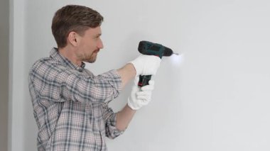 Gri gömlekli ve beyaz eldivenli orta yaşlı işçi, ev yenileme çalışmaları sırasında kablosuz matkapla duvarda delik açıyor. Dikey görünüm.