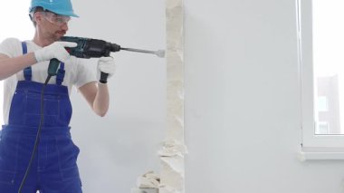 Mavi tulumlu ve koruyucu beyaz eldivenli bir inşaat işçisi, beyaz duvarı döner çekiç matkabıyla yıkıyor, toz üretiyor, yatay görüşü kapatıyor. Yenileme konsepti.