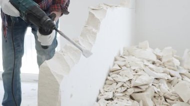 Kırmızı kareli kotlar ve koruyucu eldivenler giyen kimliği belirsiz erkek inşaat işçisi, döner çekiç matkabıyla beyaz duvarı yıkıyor, yerde enkaz oluşturuyor, manzarayı kapatıyor. Yenileme.