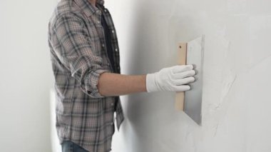 Bilinmeyen erkek inşaat işçisi koruyucu eldivenler ve ekose gömlek giyiyor. Tadilat projesi sırasında profesyonel alçıpan bantlama bıçağını kullanarak duvarın yüzeyine sorunsuzca alçı yayıyor..