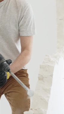 Beyaz tişörtlü ve koruyucu eldivenli, kimliği belirsiz bir inşaat işçisi, beyaz duvarı döner çekiç matkapıyla yıkıyor, yerde enkaz oluşturuyor, dikey yakın görüş açısında. Yenileme.