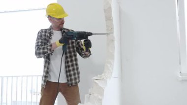 Bej kareli gömlek, sarı başlık ve koruyucu eldiven giyen inşaat işçisi, dönen çekiç matkabıyla beyaz duvarı yıkıyor ve orta uzunlukta toz üretiyor. Yenileme konsepti