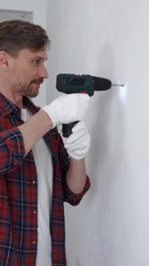 Kırmızı tişört ve beyaz eldiven giyen orta yaşlı erkek işçi, ev yenileme çalışmaları sırasında kablosuz matkapla duvarda delik açıyor. Dikey görünüm.