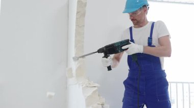 Mavi tulumlu ve koruyucu beyaz eldivenli bir inşaat işçisi, beyaz duvarı döner çekiç matkabıyla yıkıyor, toz üretiyor, yatay görüşü kapatıyor. Yenileme konsepti.