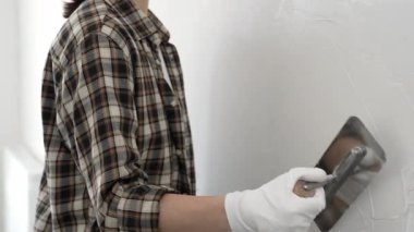 Koruyucu eldiven ve kareli tişört giyen kimliği belirsiz kadın inşaat işçisi, beyaz duvarda ıslak alçıyı düzleştiriyor ve iç dekorasyon sırasında profesyonel malayla malzeme yayıyor.