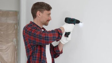 Kırmızı tişört ve beyaz eldiven giyen orta yaşlı erkek işçi, ev yenileme çalışmaları sırasında kablosuz matkapla duvarda delik açıyor. Dikey görünüm.