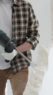 Tanınmayan bej kareli gömlek ve koruyucu eldivenler giyen erkek inşaat işçisi, döner çekiç matkabıyla beyaz duvarı yıkıyor, toz üretiyor, yakın görüş sağlıyor. Yenileme konsepti.