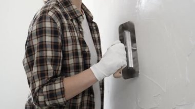 Koruyucu eldiven ve kareli tişört giyen kimliği belirsiz kadın inşaat işçisi, beyaz duvarda ıslak alçıyı düzleştiriyor ve iç dekorasyon sırasında profesyonel malayla malzeme yayıyor.