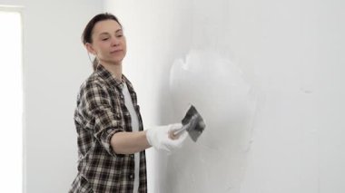 Koruyucu eldiven ve kareli gömlek giyen kadın, iç dekorasyon çalışmalarını boyamadan önce hasarlı bölgeleri yumuşatmak için beyaz duvar yüzeyine macun sürüyor..