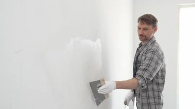 Koruyucu eldiven ve ekose gömlek giyen erkek inşaat işçisi, tadilat projesi sırasında profesyonel alçı levha bantlama bıçağı kullanarak duvarın yüzeyine sorunsuzca alçı yayıyor. Dikey görünüm.