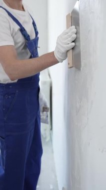 Koruyucu eldivenler ve mavi inşaat tulumları giyen erkek inşaat işçisi, tadilat projesi sırasında profesyonel alçı levha bantlı bıçak kullanarak duvarlara sorunsuzca alçı yayıyor. Portre