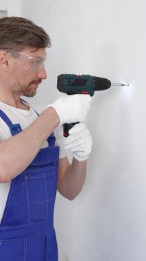 Mavi üniformalı profesyonel işçi, güvenlik eldivenleri, tadilat sırasında beyaz duvarda delik açmak için kablosuz matkap kullanıyor. Dikey portre görünümü.