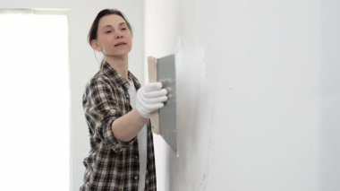 Kareli gömlek giyen kadın inşaat işçisi, beyaz duvar yüzeyini düzleştiriyor, profesyonel spatula tekniği ile macunu uyguluyor, iç dekorasyon konusunda yetenekli..