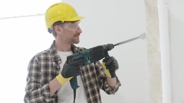 Bej kareli gömlek, sarı başlık ve koruyucu eldiven giyen inşaat işçisi, döner çekiç matkabıyla beyaz duvarı yıkıyor, toz ve portre görüntüsü yaratıyor. Yenileme konsepti.