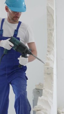 Mavi tulumlu ve koruyucu beyaz eldivenli bir inşaat işçisi, beyaz duvarı döner çekiç matkabıyla yıkıyor, toz üretiyor, yatay görüşü kapatıyor. Yenileme konsepti.