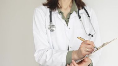 Stetoskop ve yeşil bluzun üzerine beyaz önlük giyen kimliği belirsiz bir kadın doktor tahta renkli bir panoya tıbbi uzmanlığı ve hasta bakımını temsil eden bir yazı yazıyor. Tıp konsepti