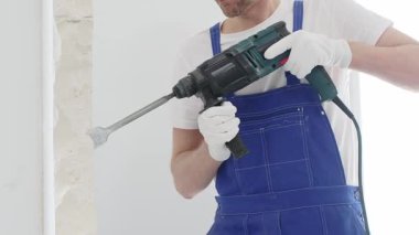 Mavi tulumlu ve koruyucu beyaz eldivenli tanımlanamayan erkek inşaat işçisi, beyaz duvarı döner çekiç matkapıyla yıkıyor, toz üretiyor ve yatay görüşü kapatıyor. Yenileme