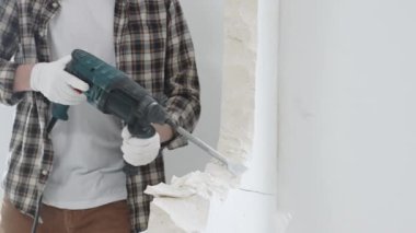 Tanınmayan bej kareli gömlek ve koruyucu eldivenler giyen erkek inşaat işçisi, döner çekiç matkabıyla beyaz duvarı yıkıyor, toz üretiyor, yakın görüş sağlıyor. Yenileme konsepti.