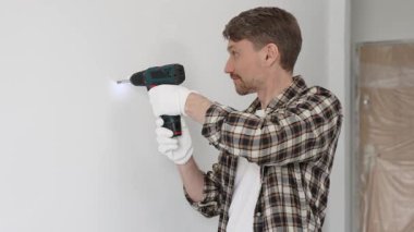 İşaretli gömlek ve güvenlik eldiveni giyen erkek inşaat işçisi, kablosuz tornavida kullanarak duvara vida sokuyor ve dikkatli bir ev tadilatı yapıyor. Dikey görünüm.